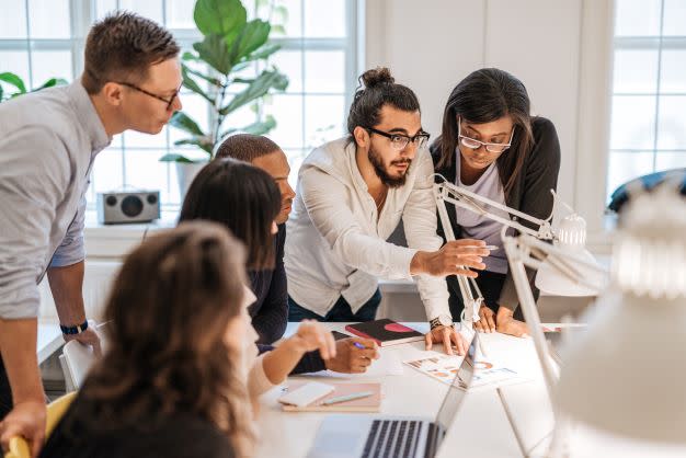 Team of six co-workers working together on a project. They are standing over a table in a modern office or workspace and discussing while referring to a piece of paper showing a series of graphs.