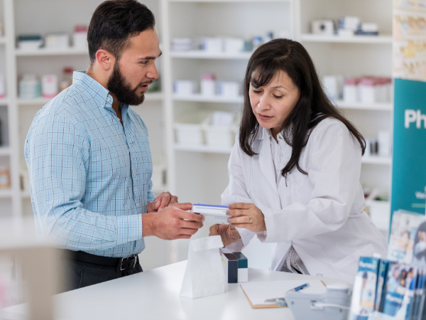 A pharmacist in a white lab coat is speaking to a man while they are both holding and looking at a box of medication.