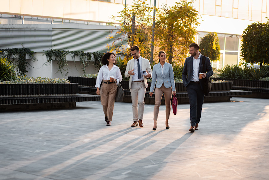 Four business professionals walking together outside a modern office building while talking and holding coffee cups.