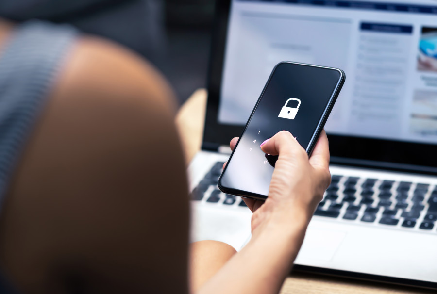 Woman sitting at an open laptop using her mobile phone. There is a digital lock icon on her phone screen, representing data security and digital privacy.