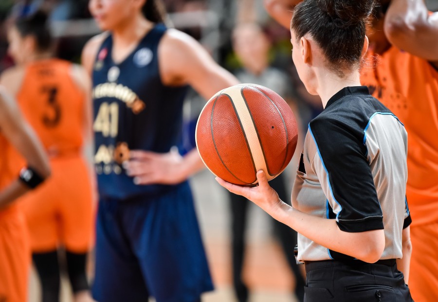 A women's basketball team on the court. In the foreground is a referee holding a basketball; she has her back to the viewer.