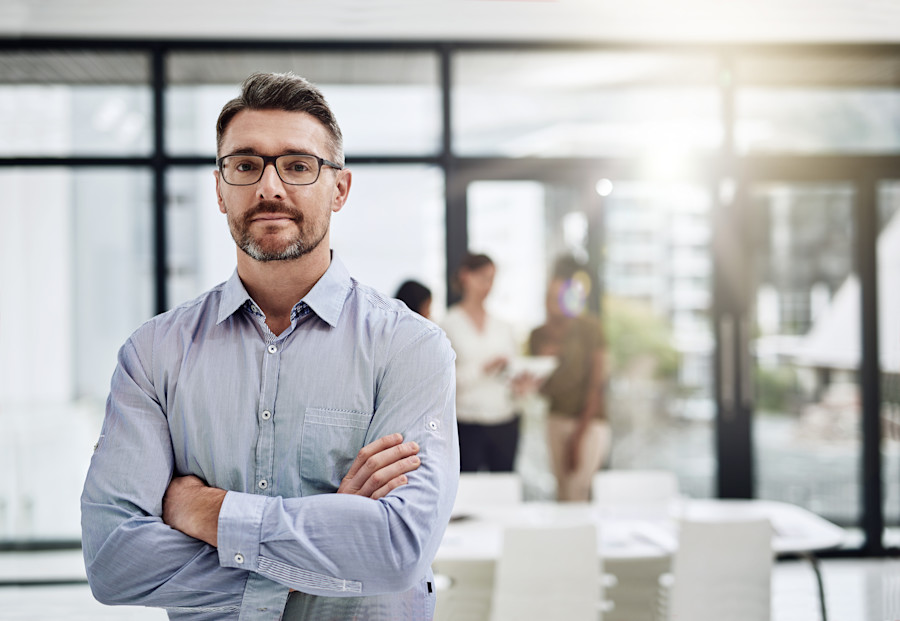 Professional man standing with his arms crossed. Behind him is an office environment with a small group of colleagues gathered.