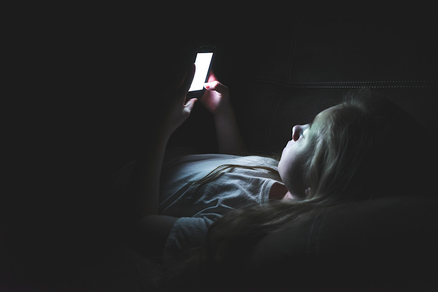 A young girl or child lying in bed in the dark, reading the internet on her phone. 
