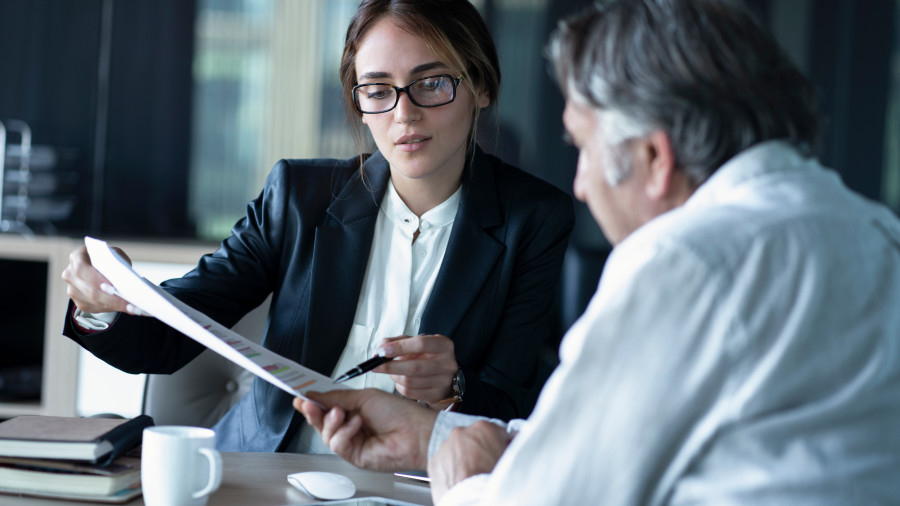 Female lawyer or advisor sitting at a desk in front of a male client. She is showing him a report or piece of writing on a paper and talking him through it.