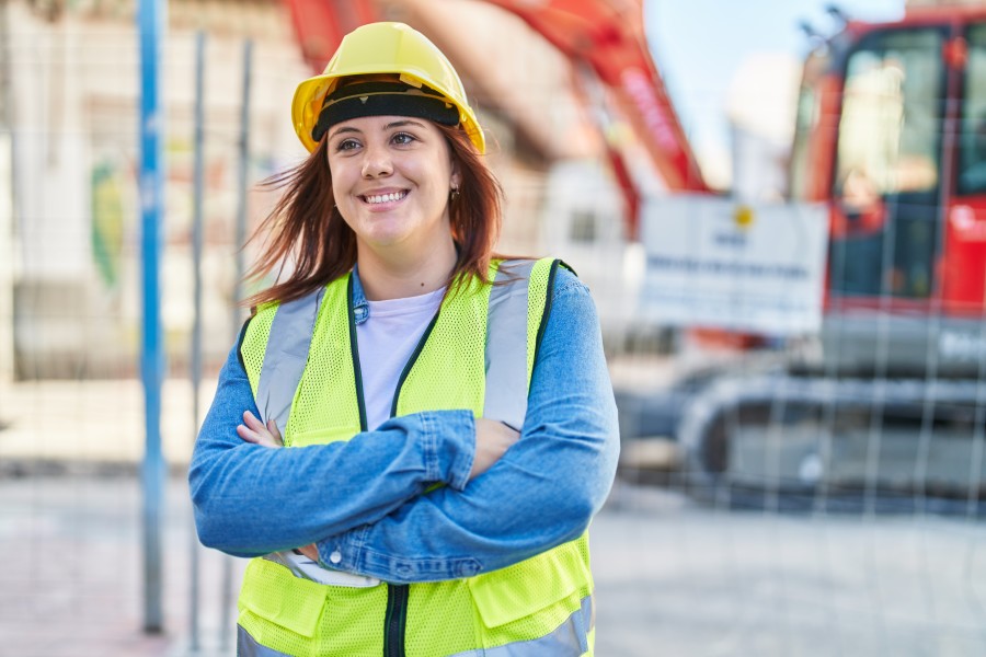 Female worker in yellow high-vis vest and hard hat standing in front of a construction or work site with her arms crossed.