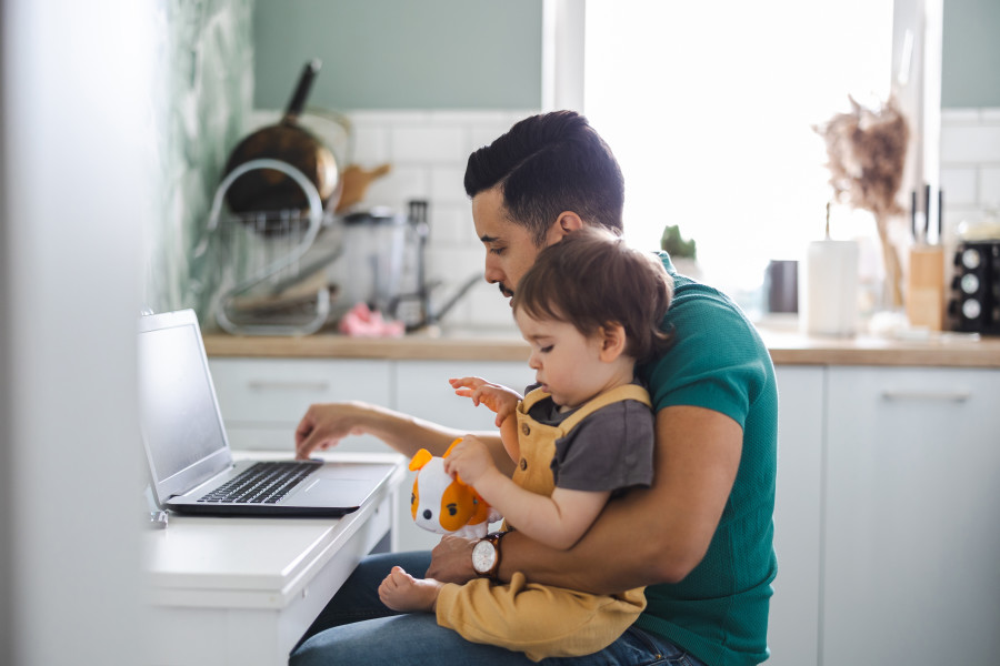 Father with a child on his lap, sitting at a desk and working on a laptop.