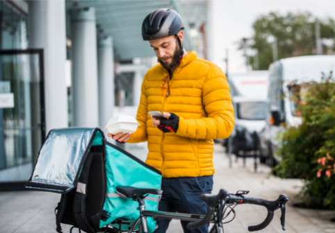Food delivery cyclist standing next to bike, holding a takeaway container and checking his phone.