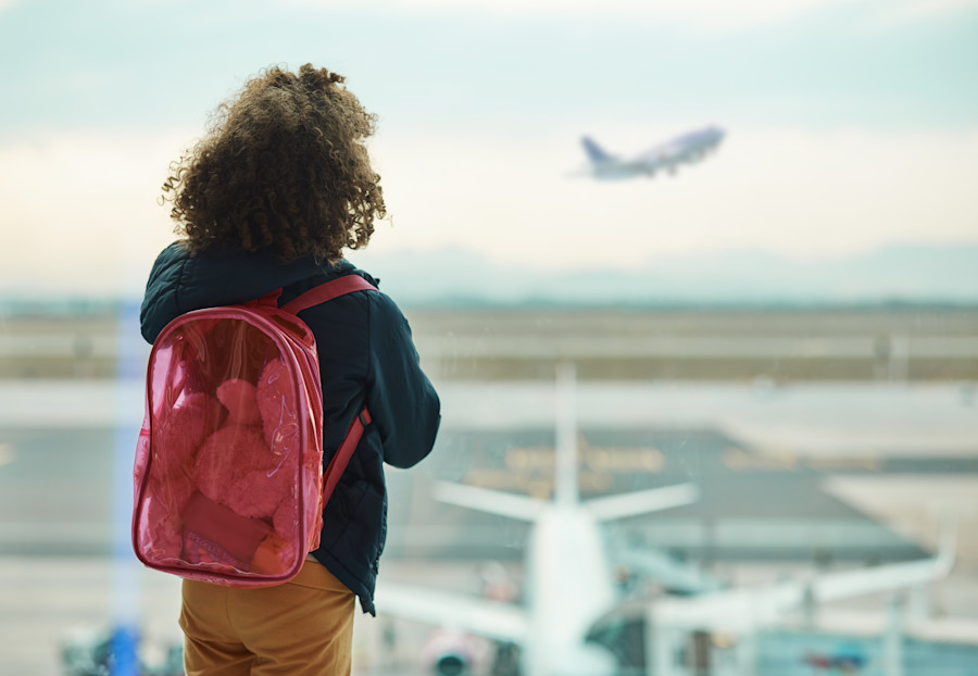 A small child wearing a backpack is standing and watching a plane take off a runway through a window at an airport. 