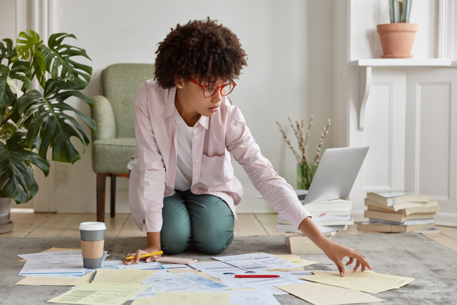 Woman kneeling on the floor of her house of apartment, looking through pages of notes and documents.