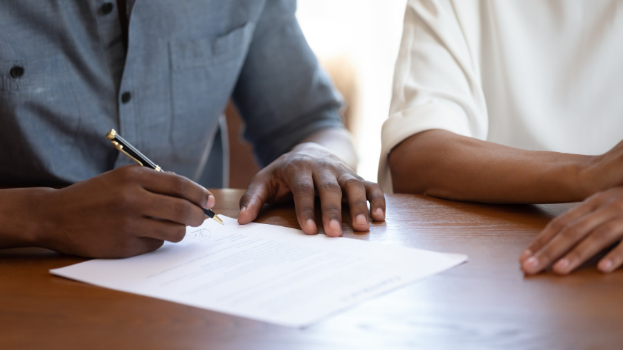 Two people sitting at a desk; one is signing a document with a pen.