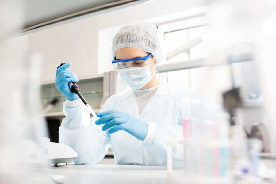 A woman wearing a mask, safety goggles and hairnet is working in a laboratory. She is using a teat pipette. 