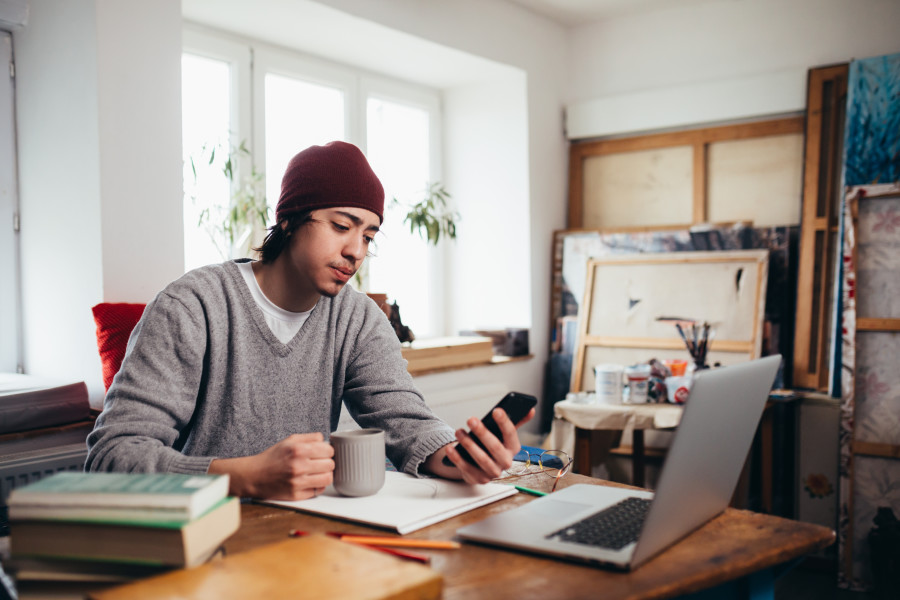 Young male worker sitting at his home office desk in front of an open laptop. He is holding a mobile phone in one hand, looking at the screen, and a mug in his other hand.