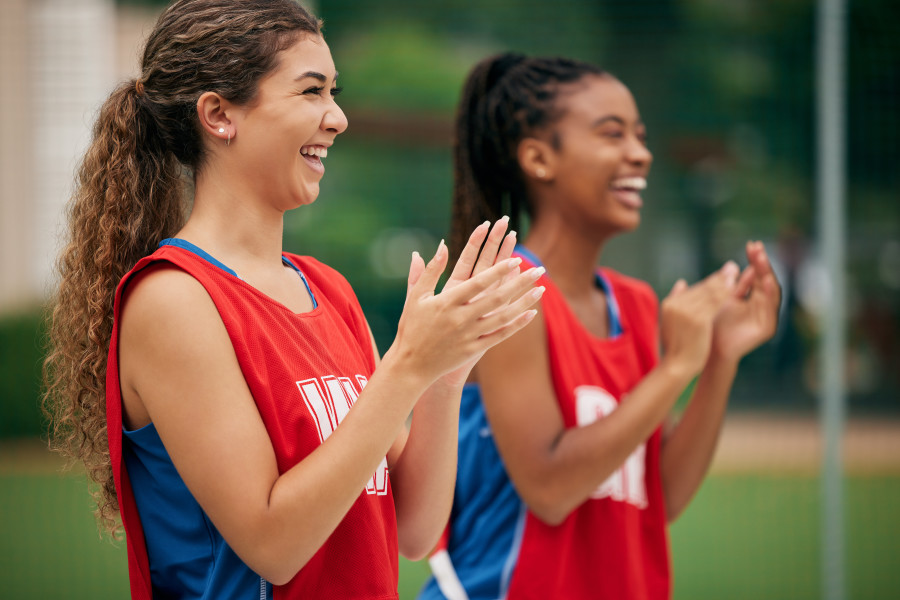 Two female netballers in red bibs standing next to each other, smiling and clapping.