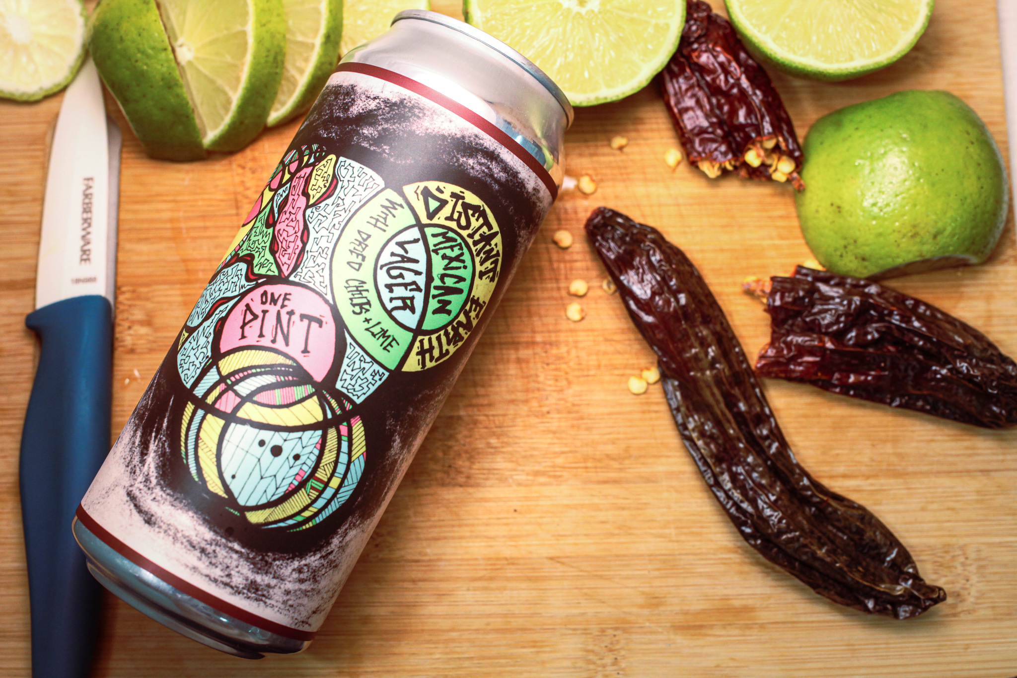 A 16oz can of mexican lager is laid down on a cutting board surrounded by dried chilis and limes