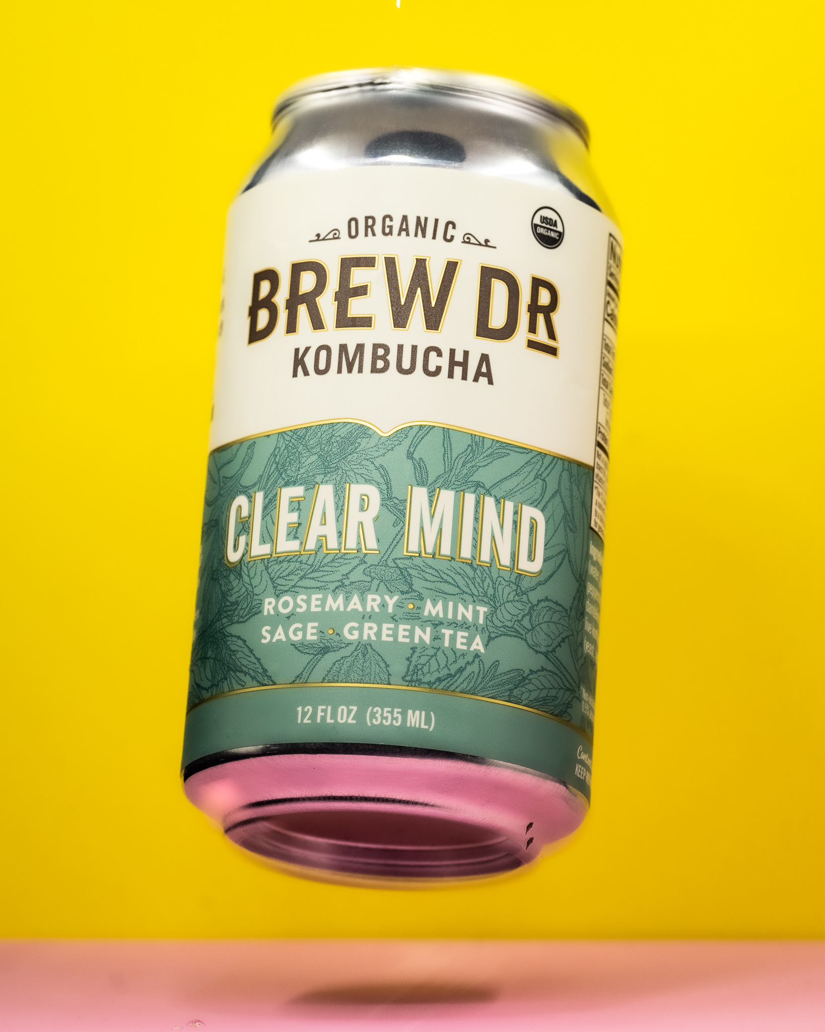 A 12 oz can  of Brew Dr. Clear Mind Kombucha, suspended above a pink floor and in front of a bright yellow background.