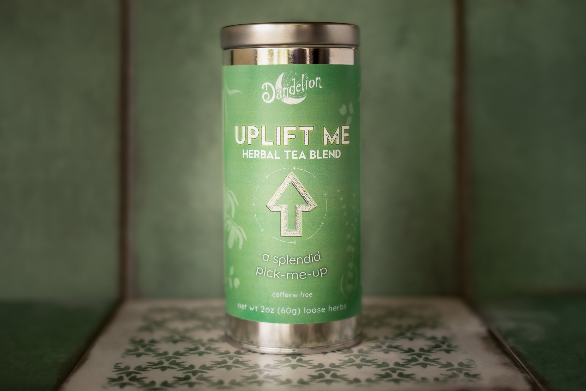A tin of Uplift Me Tea from dandelion teahouse sits on light green and white floral pattern tile. The label is mint green with yellow accents and an "UP" arrow in the middle of it.