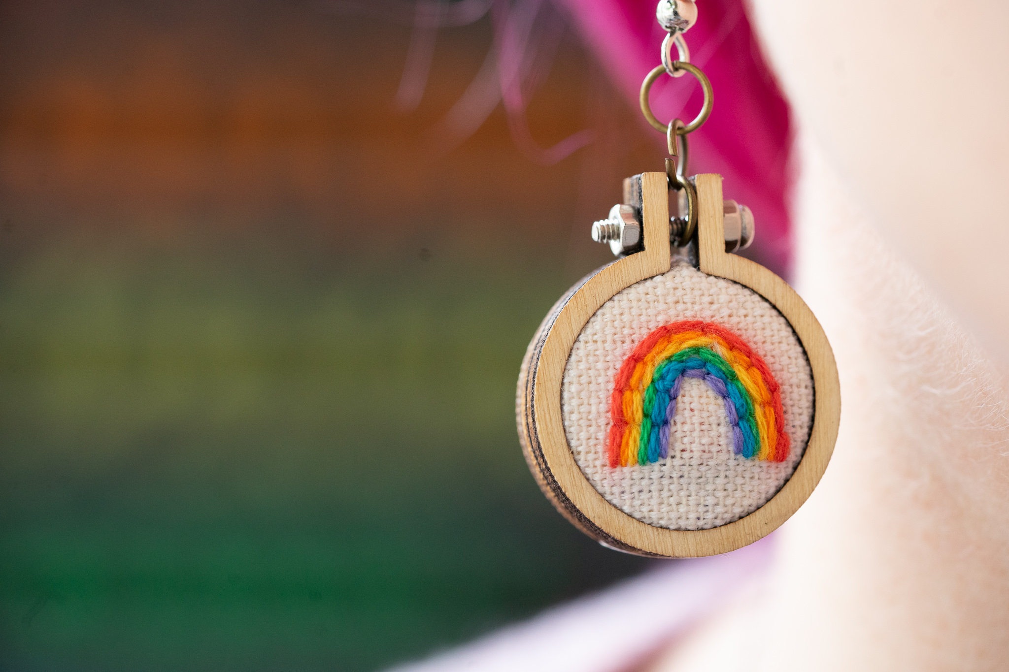 A rainbow embroidered on a 1 inch circle of white fabric is attached to a circular earring, dangling from a girl with pink hair's ear.