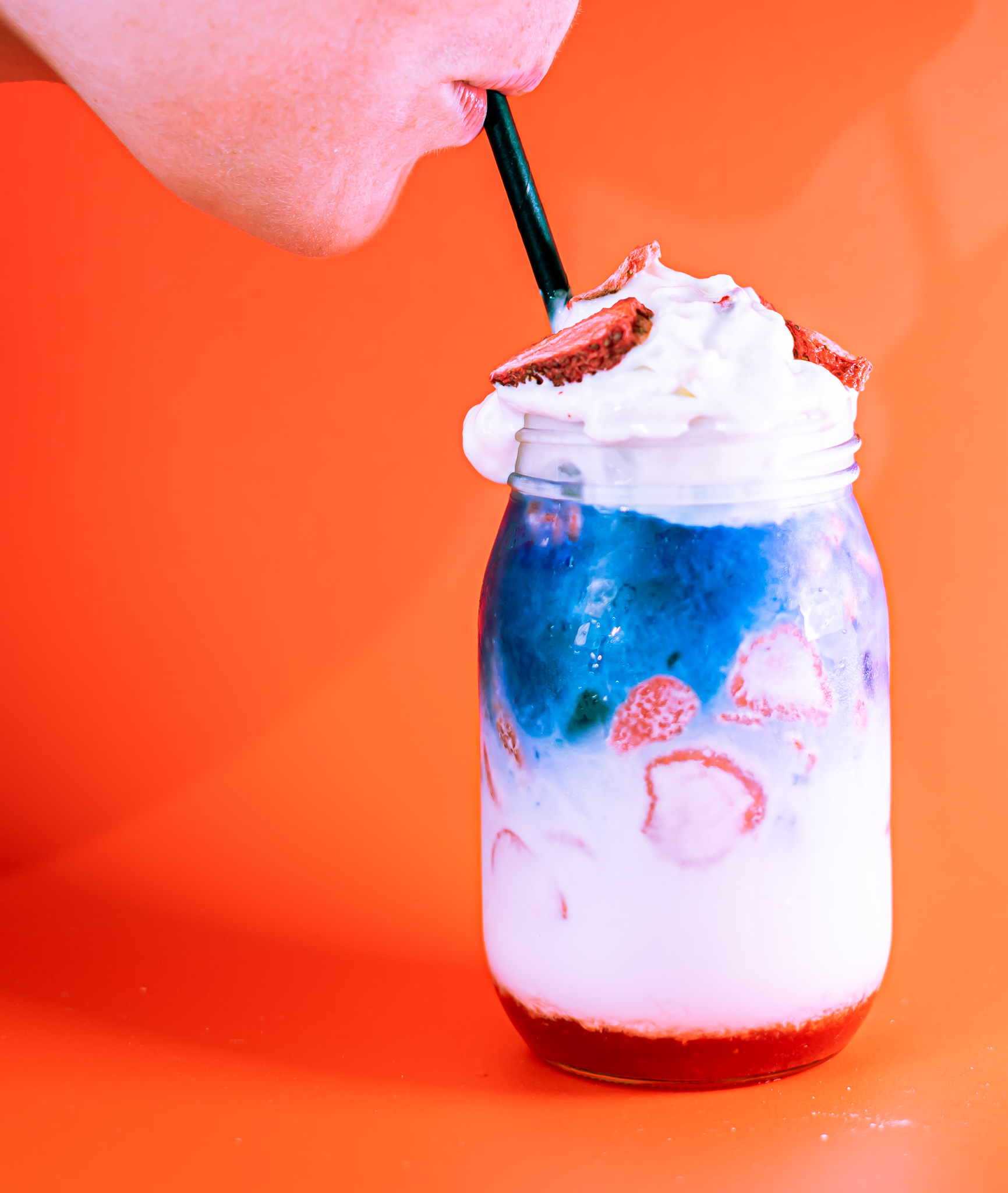 A woman sipping from a layered red white and blue tea drink in a mason jar, topped with whipped cream and dried strawberries in front of a red background.