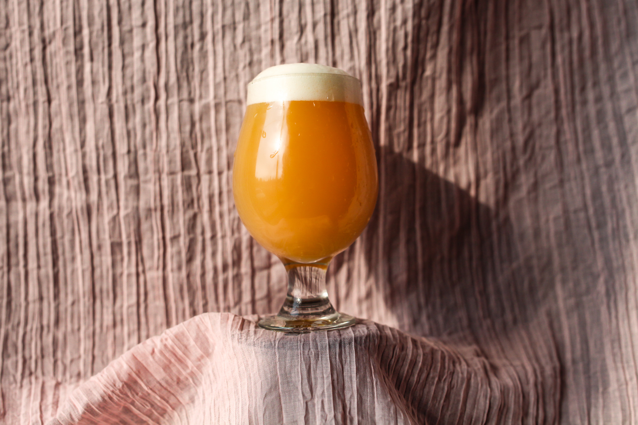 A bright orange hazy IPA in a tulip glass against a wrinkled pink lined cloth backdrop. The creases in the cloth create dramatic shadows trending  from left to right.