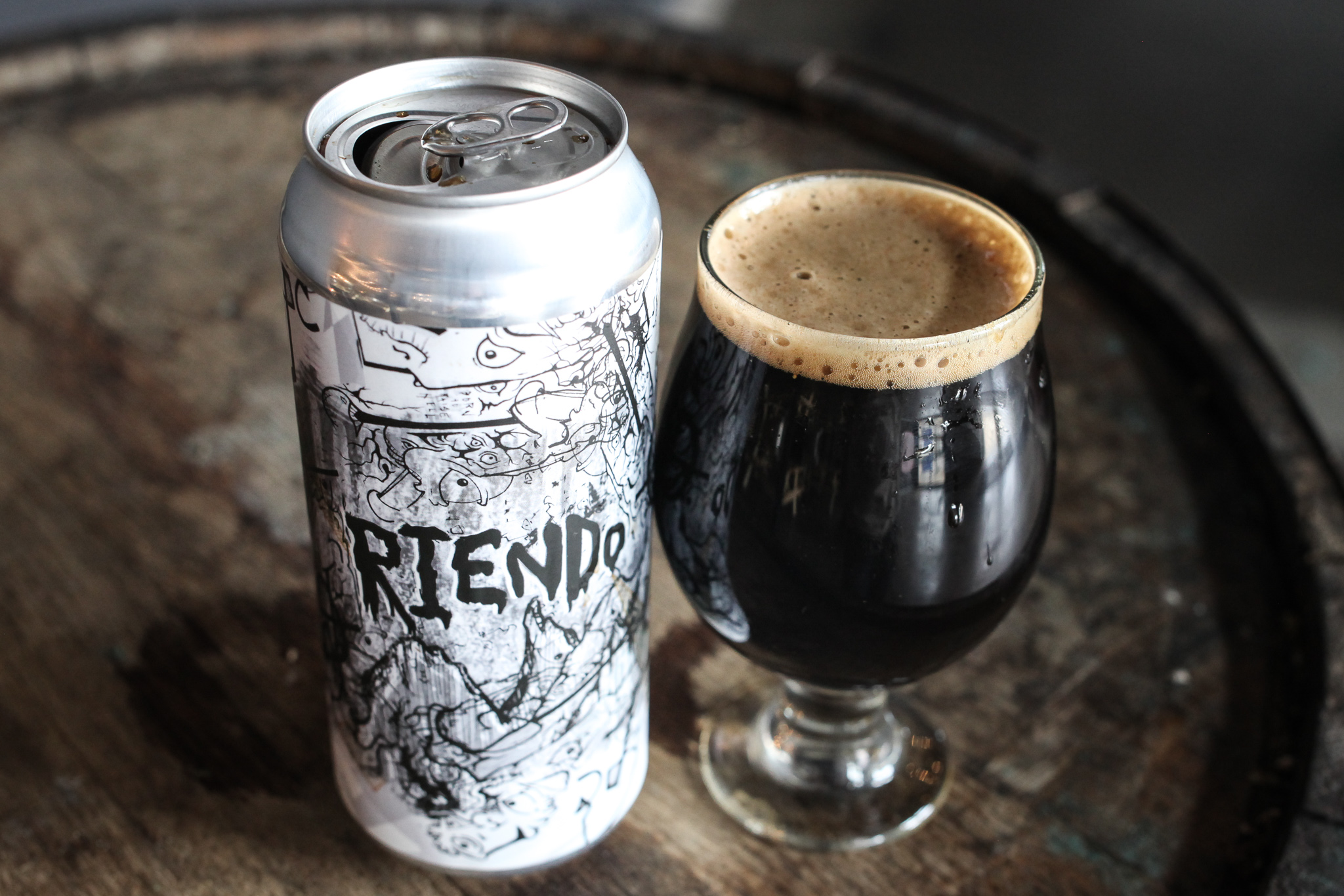 A 16oz can of beer with a black and white label reading "Riendo" next to a tulip shaped glass of a dark imperial stout