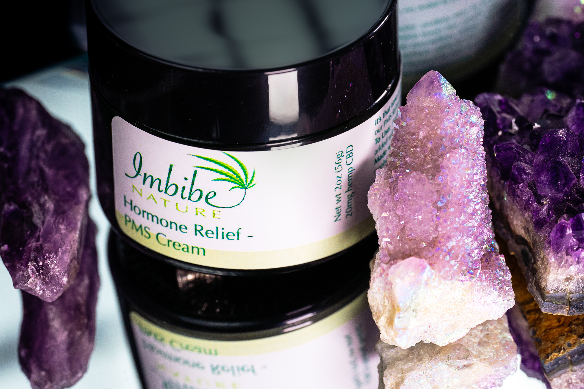 A jar of Imbibe Nature Hormone Relief PMS Cream surrounded by a variety of light and dark purple crystals.