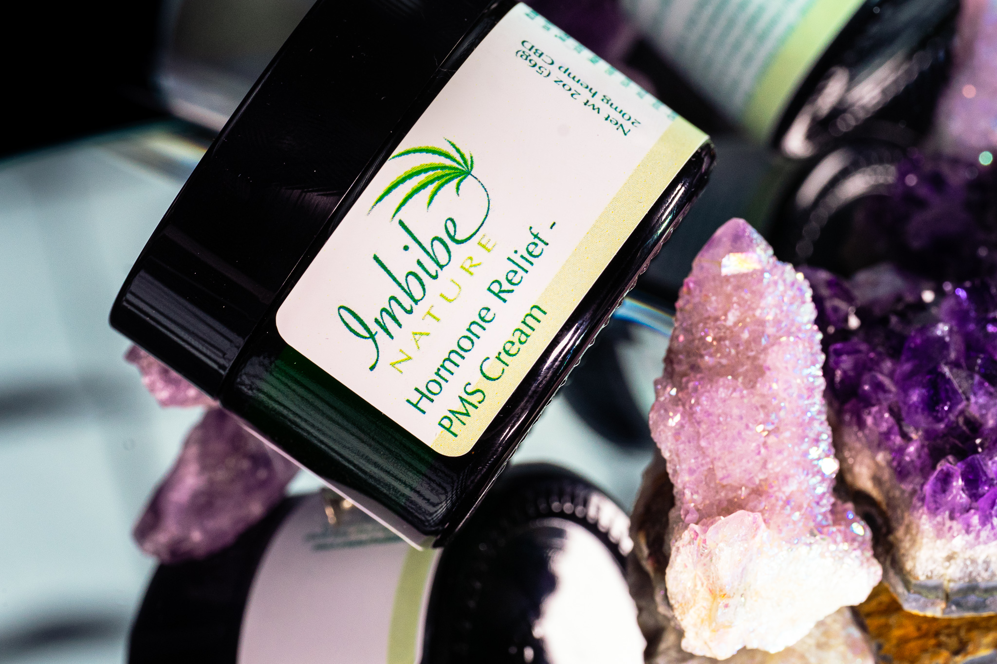 A jar of Imbibe Nature Hormone Relief PMS Cream surrounded by a variety of light and dark purple crystals.
