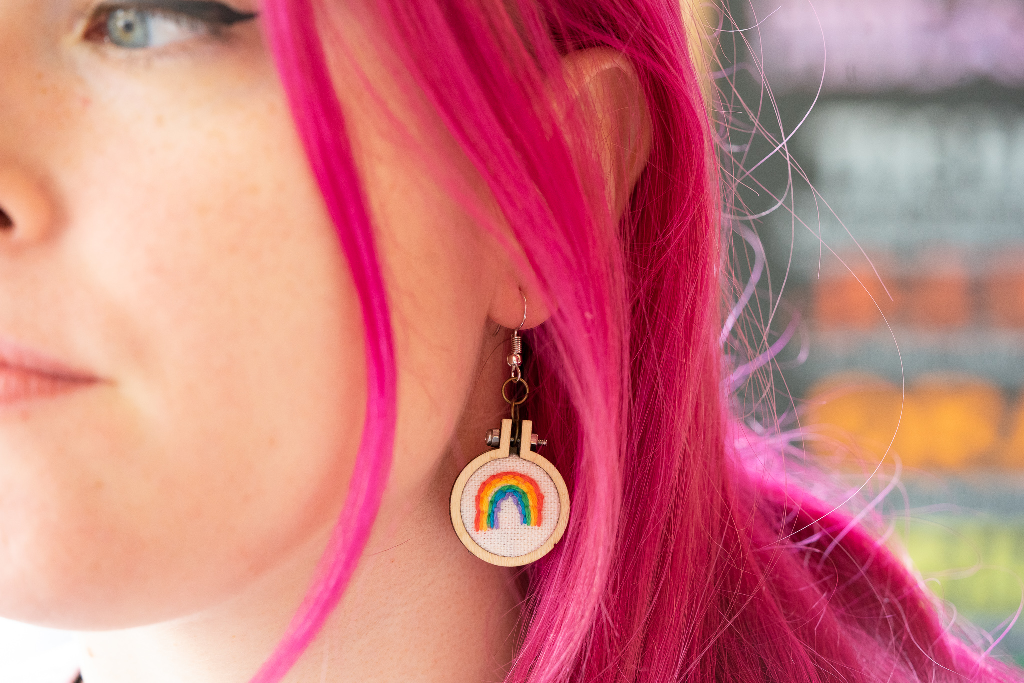 A rainbow embroidered on a 1 inch circle of white fabric is attached to a circular earring, dangling from a girl with pink hair's ear.