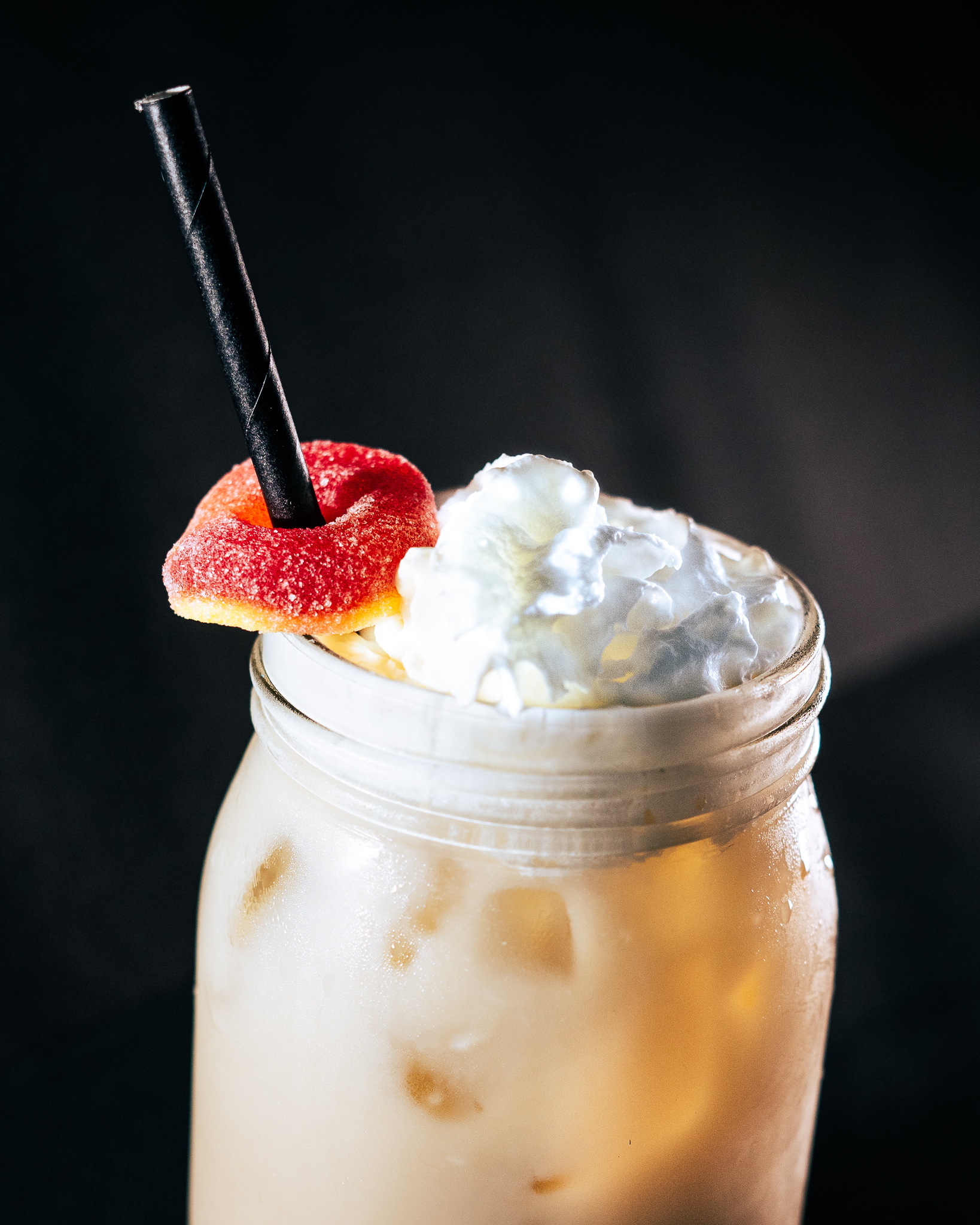 A cream colored tea drink in a mason jar sits against a black background. It's topped with whipped cream, and a candy peach ring around a black straw