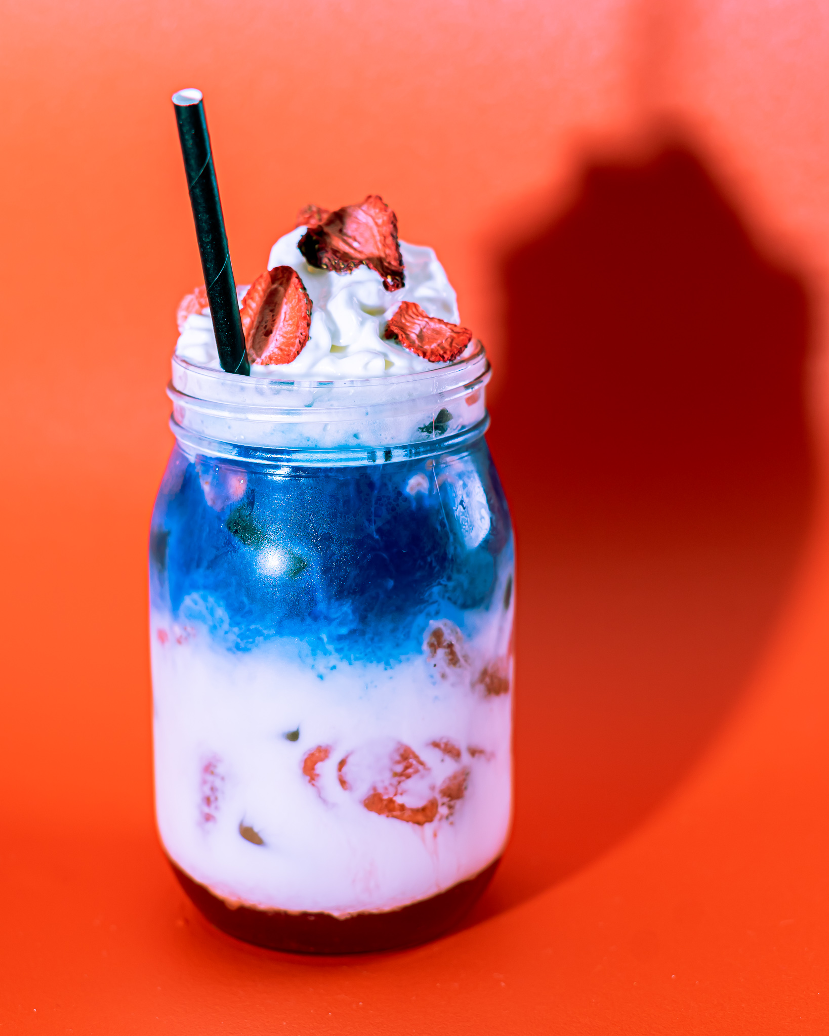 A layered red white and blue tea drink in a mason jar, topped with whipped cream and dried strawberries in front of a red background.