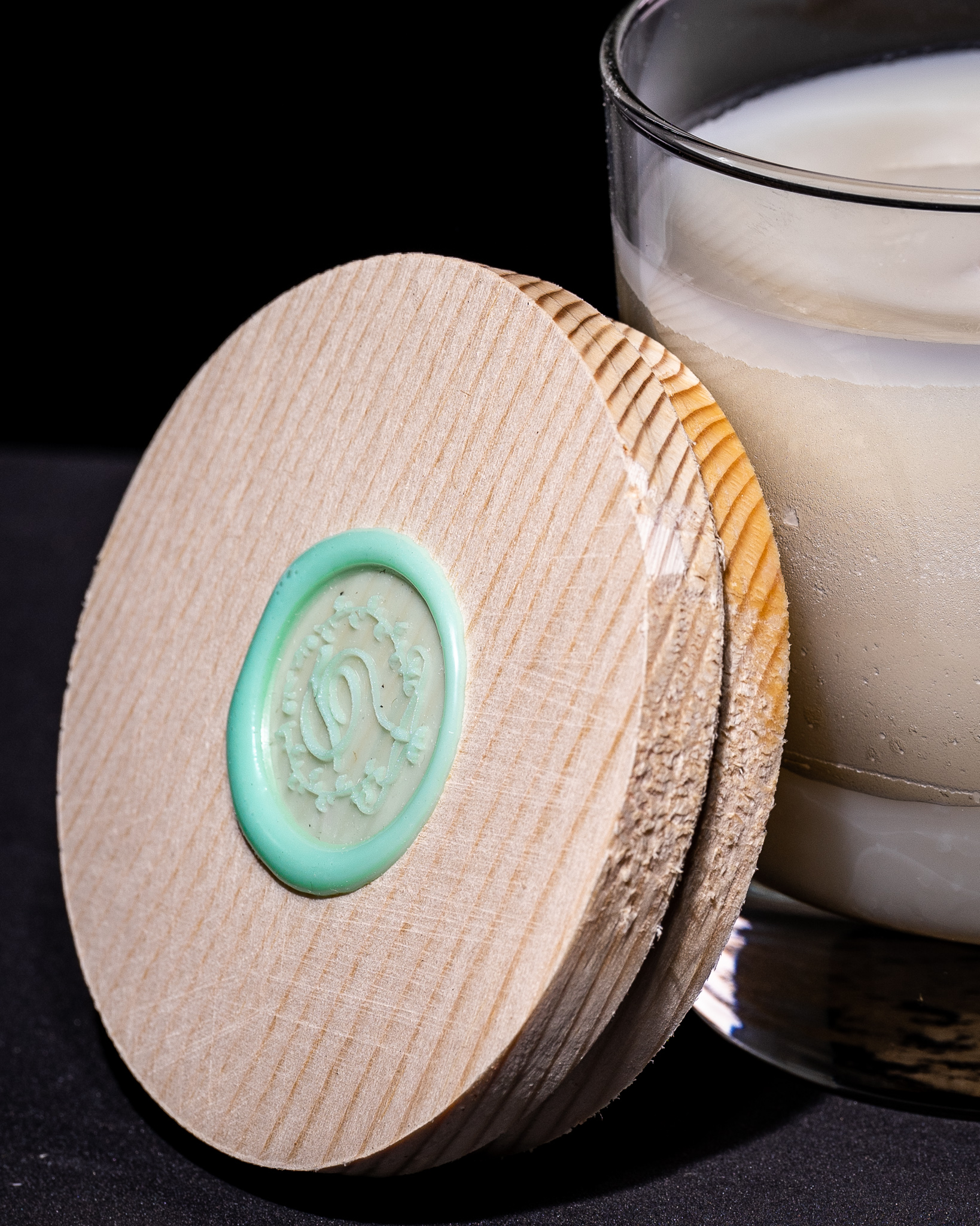 A light wooden circular lid propped up against a lilac colored candle. There is a turquoise pressed wax emblem on the center of the lid.