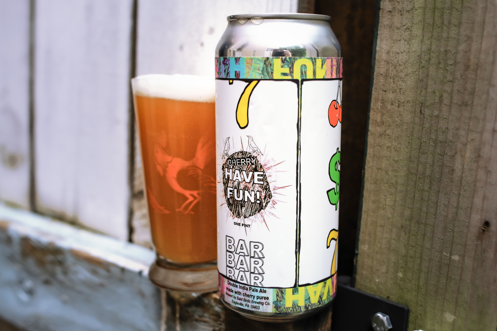 A 16 ounce beer can with a label displaying the title "Have fun" and drawings of a casino slot machine sits next to a glass of hazy IPA along a wooden fence.
