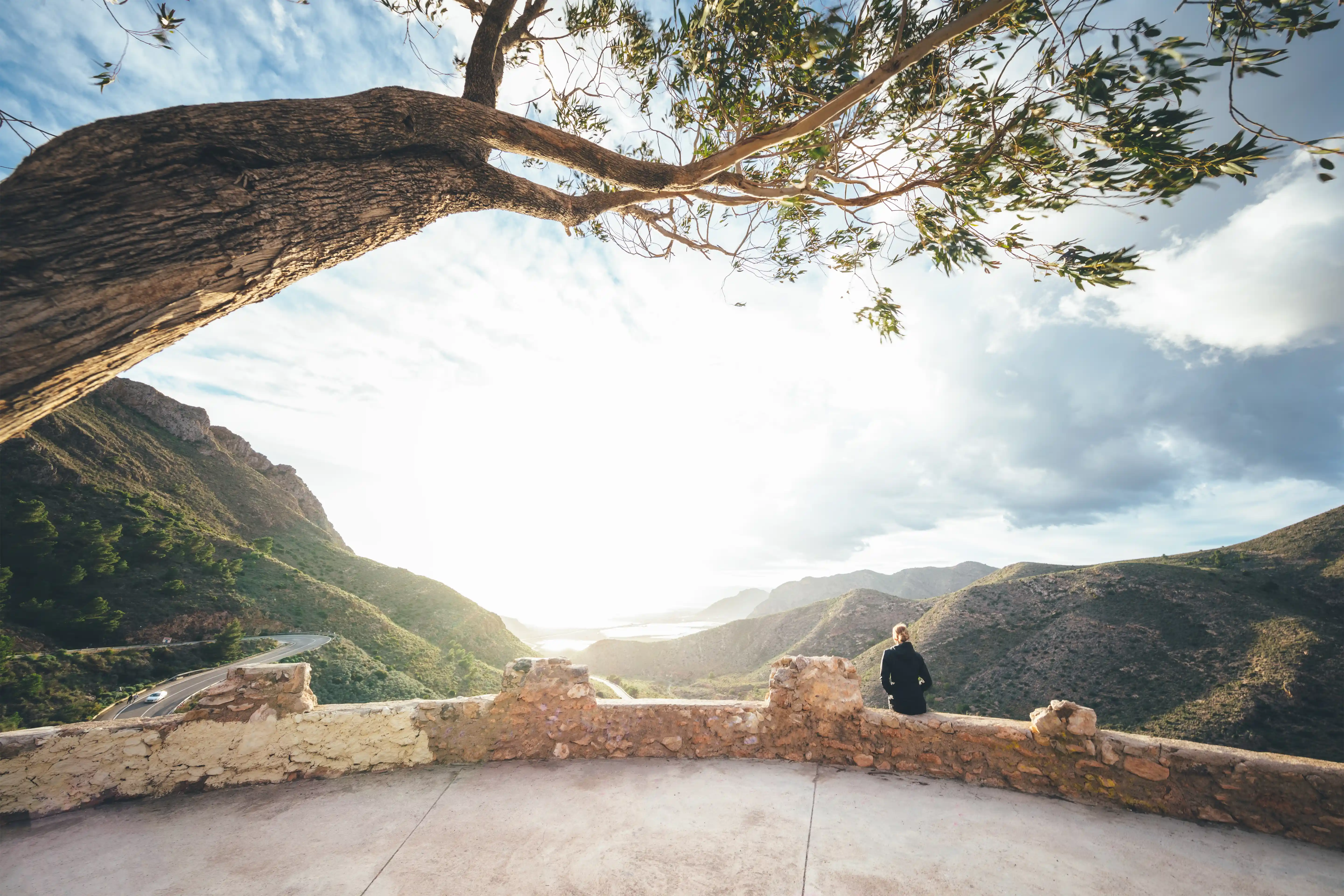 Woman sitting on the wall under the tree looking at mountain landscape in Cartagena, province of Murcia, Spain.
