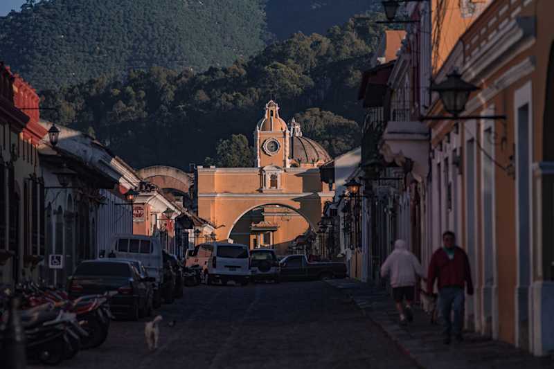 the sun setting on a colonial street with tree lined hills in the background. antigua, guatamala.