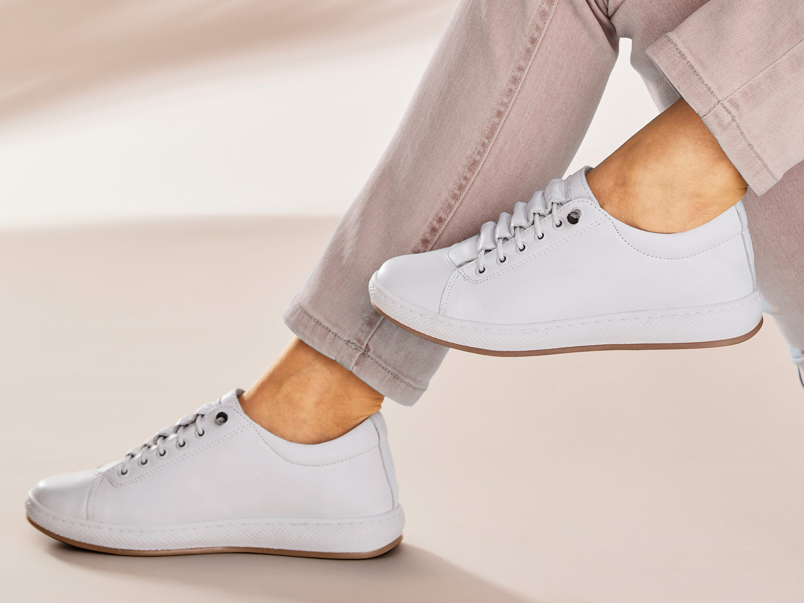 Ein Model trägt weiße, schlichte Sneaker und eine helle, leicht hochgekrempelte Hose. Der Hintergrund ist beige und minimalistisch.