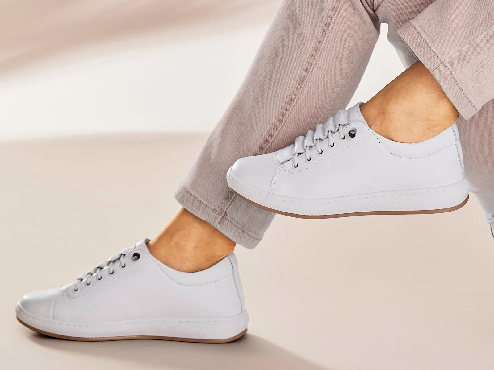 Ein Model trägt weiße, schlichte Sneaker und eine helle, leicht hochgekrempelte Hose. Der Hintergrund ist beige und minimalistisch.