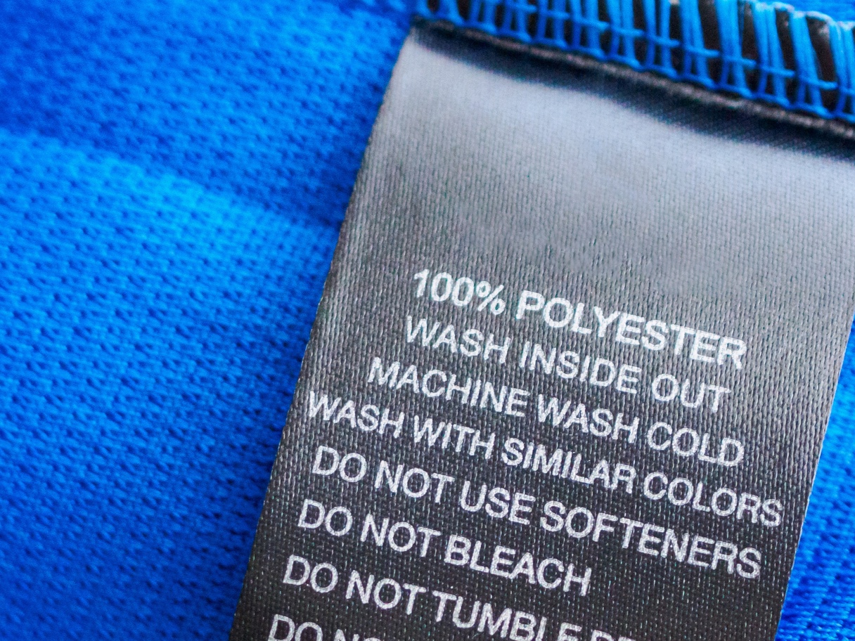 Pflegeetikett aus schwarzem Stoff auf blauem Textil, zeigt Waschhinweise für 100% Polyester.