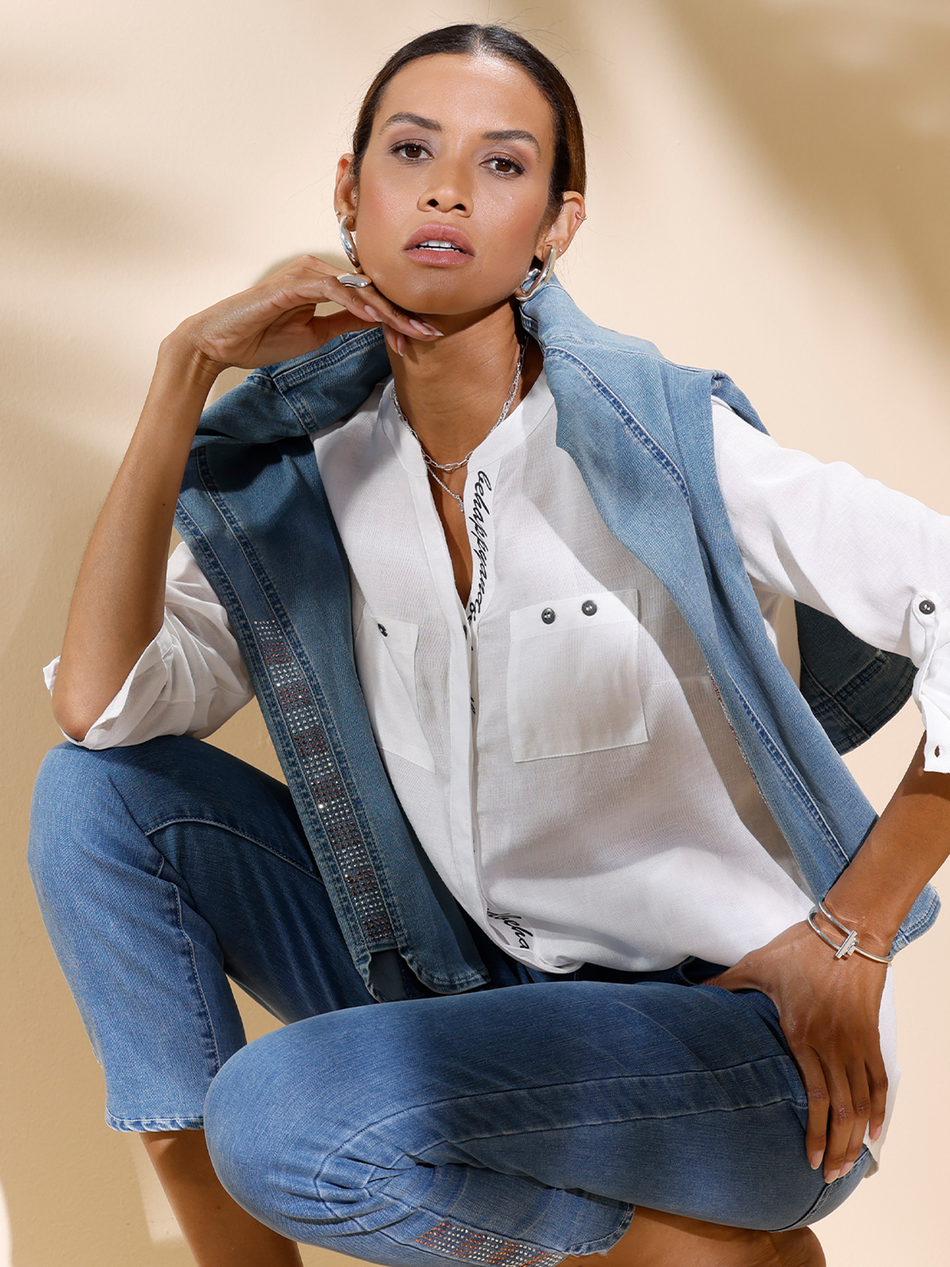 Ein Model sitzt lässig vor hellem Hintergrund, trägt eine weiße Bluse, blaue Jeans und eine aufgelegte Jeansjacke