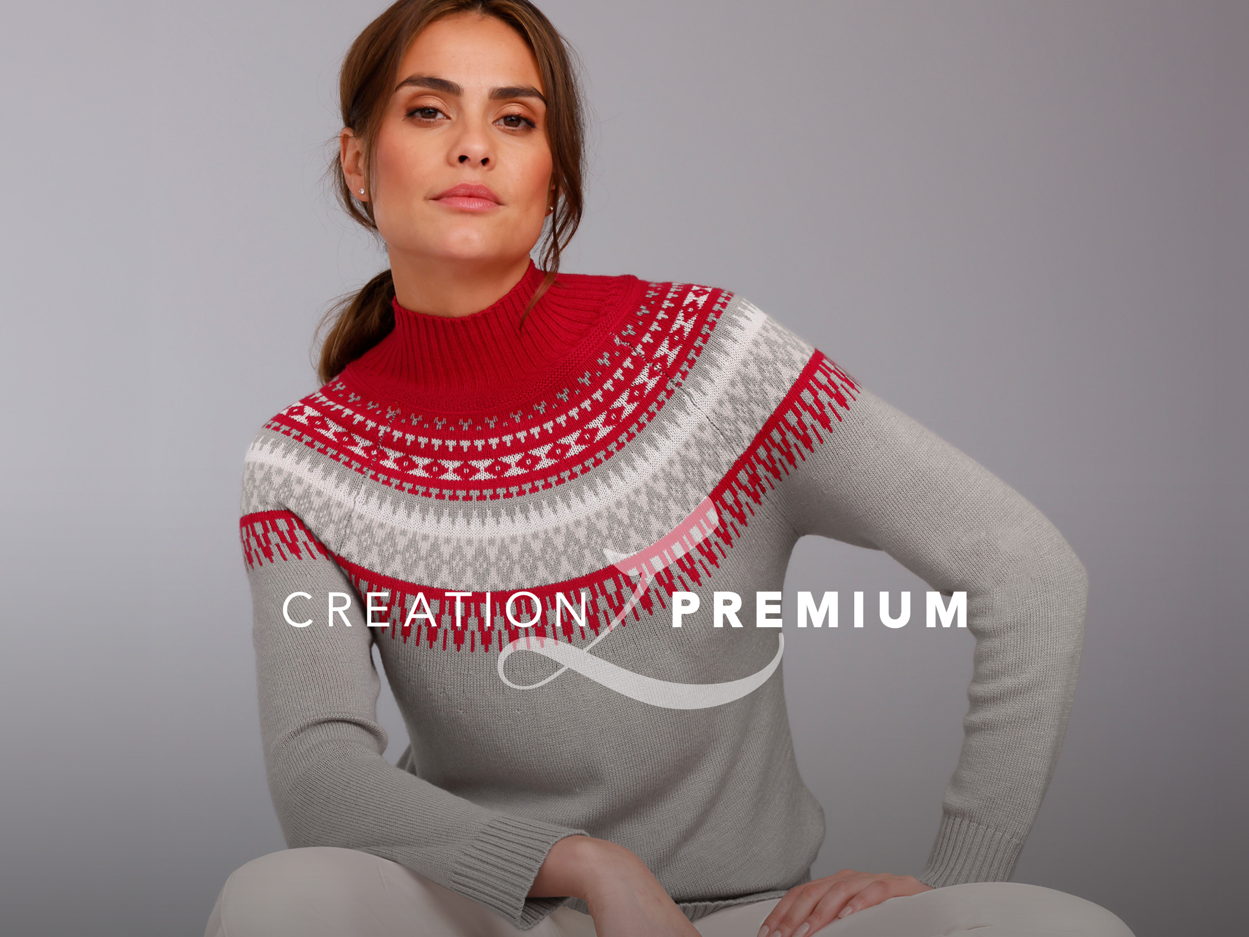 Ein Model trägt einen grauen Pullover mit rot-weißem Muster und hohem Kragen, sitzt vor neutralem Hintergrund.
