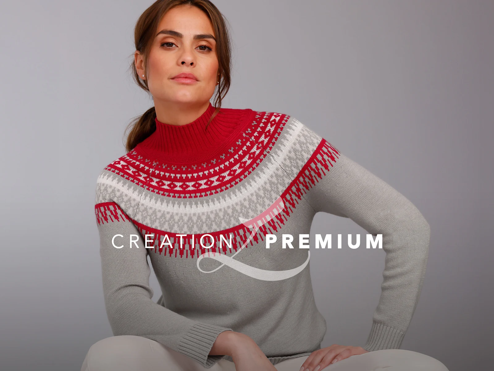 Ein Model trägt einen grauen Pullover mit rot-weißem Muster und hohem Kragen, sitzt vor neutralem Hintergrund.