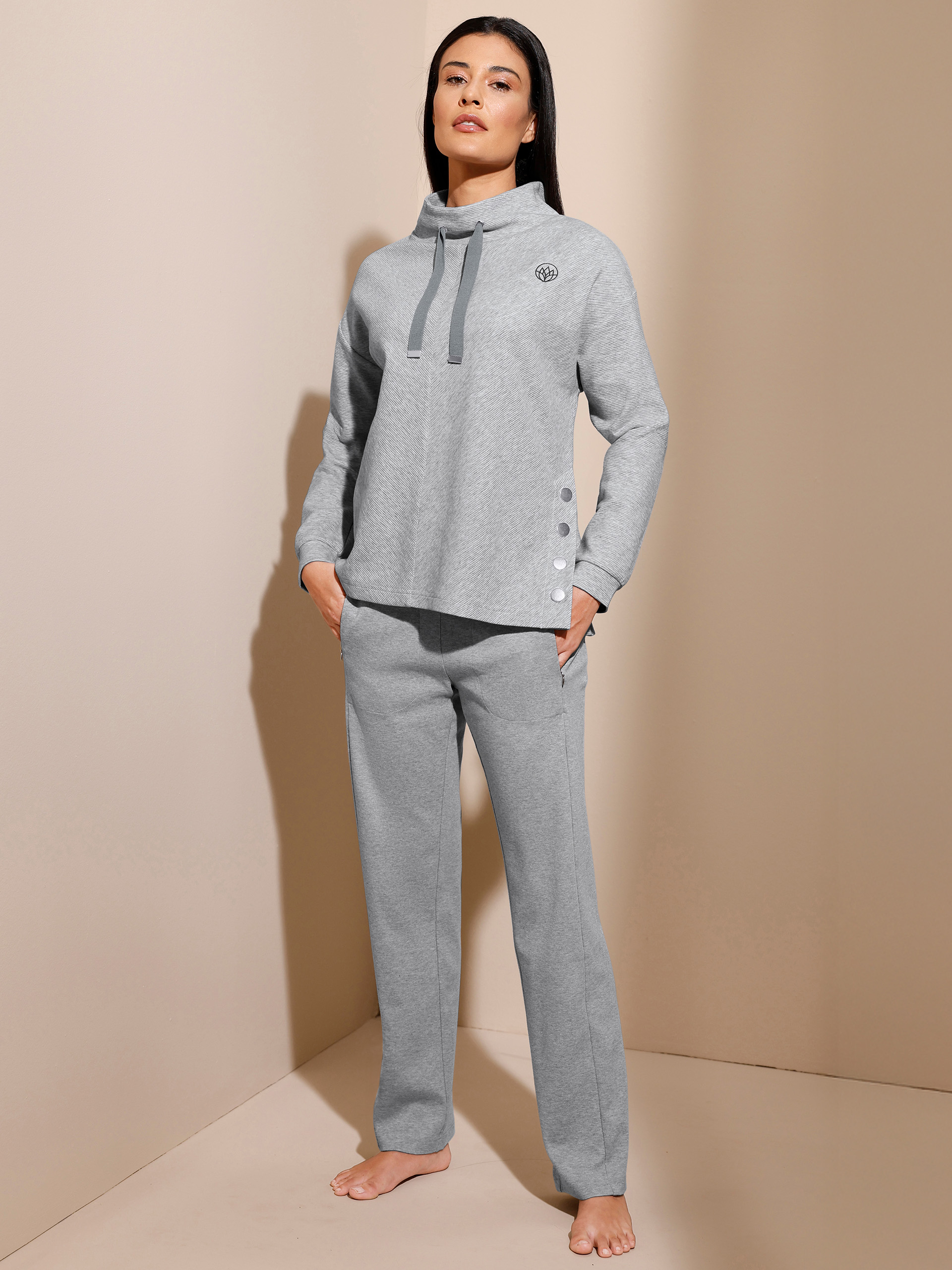 Ein Model trägt einen grauen Jogginganzug mit hohem Kragen, Kordelzug und Knöpfen an der Seite, barfuß stehend vor beigem Hintergrund.