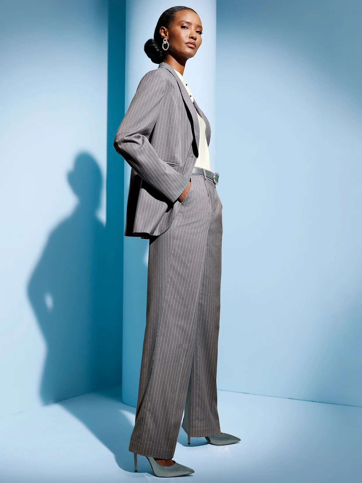 Ein Model trägt einen grauen, gestreiften Anzug mit einer weißen Bluse und hält eine silberne Clutch. Der Hintergrund ist blau. Ein Model trägt einen grauen, gestreiften Anzug mit einer weißen Bluse und hält eine silberne Clutch. Der Hintergrund ist blau.