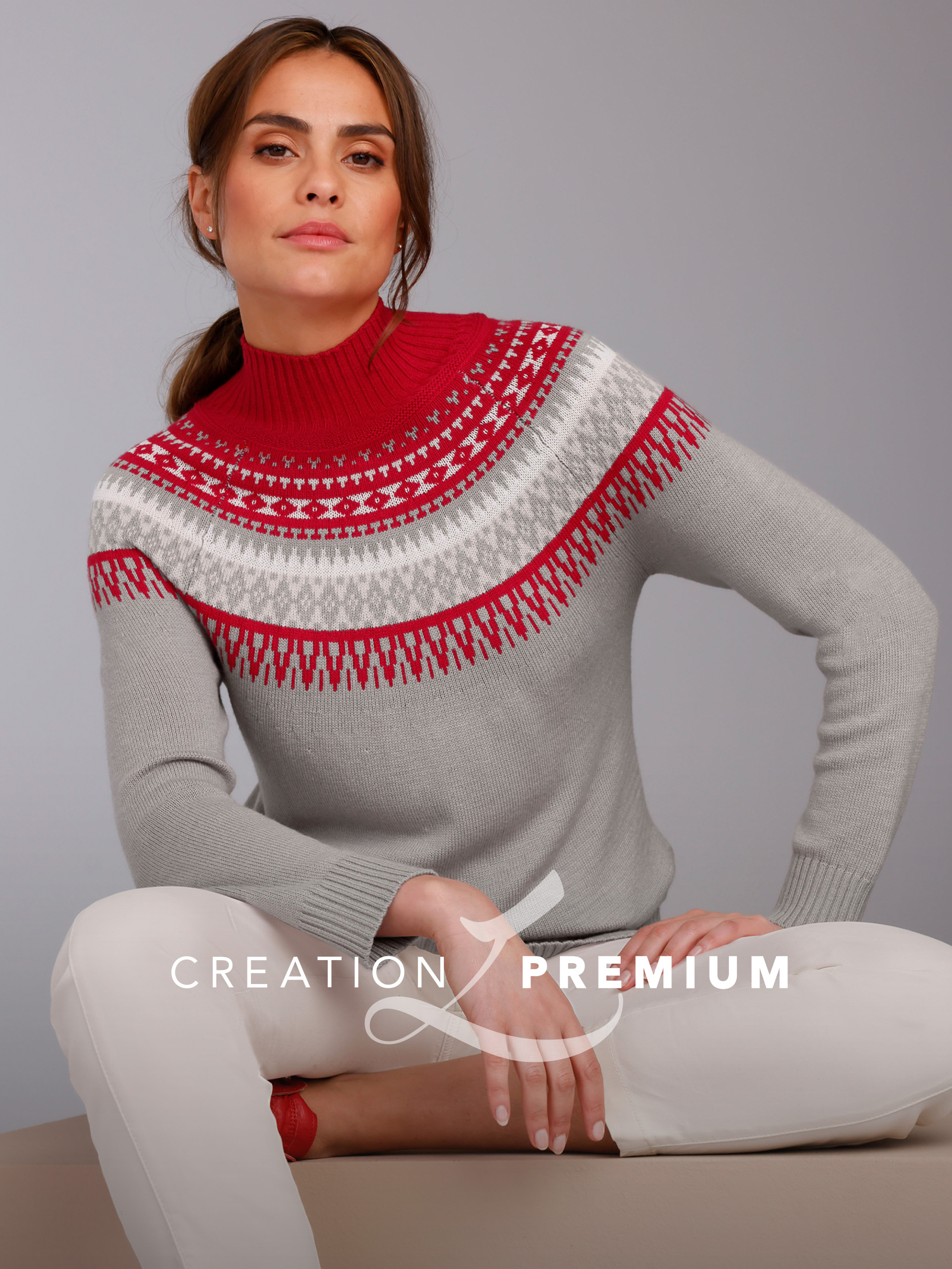 Ein Model trägt einen grauen Pullover mit rot-weißem Muster und hohem Kragen, sitzt vor neutralem Hintergrund.