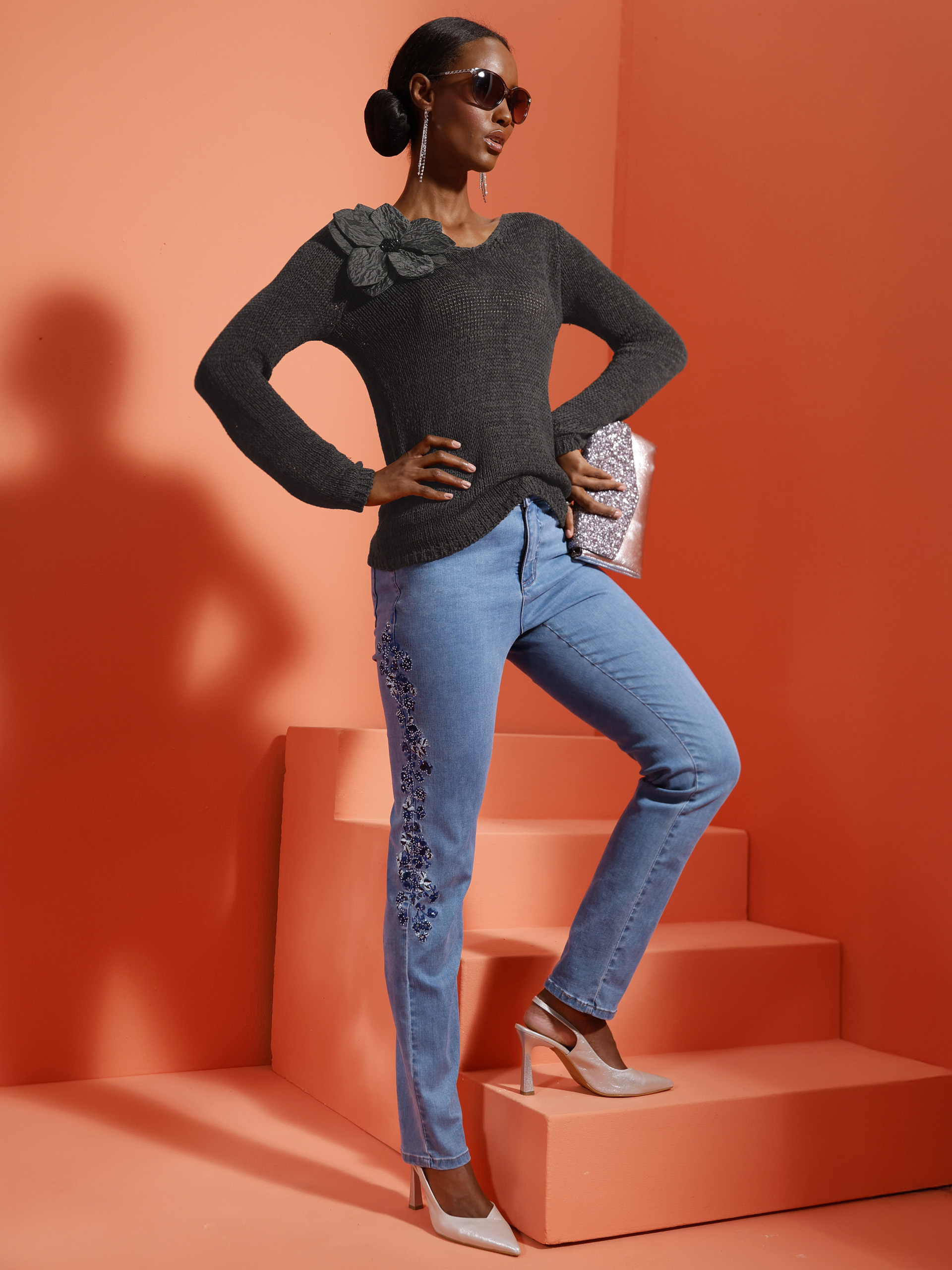 Ein Model steht auf einer Treppe vor orangem Hintergrund, trägt einen dunklen Pullover mit Blumenapplikation, bestickte Jeans und helle High Heels.