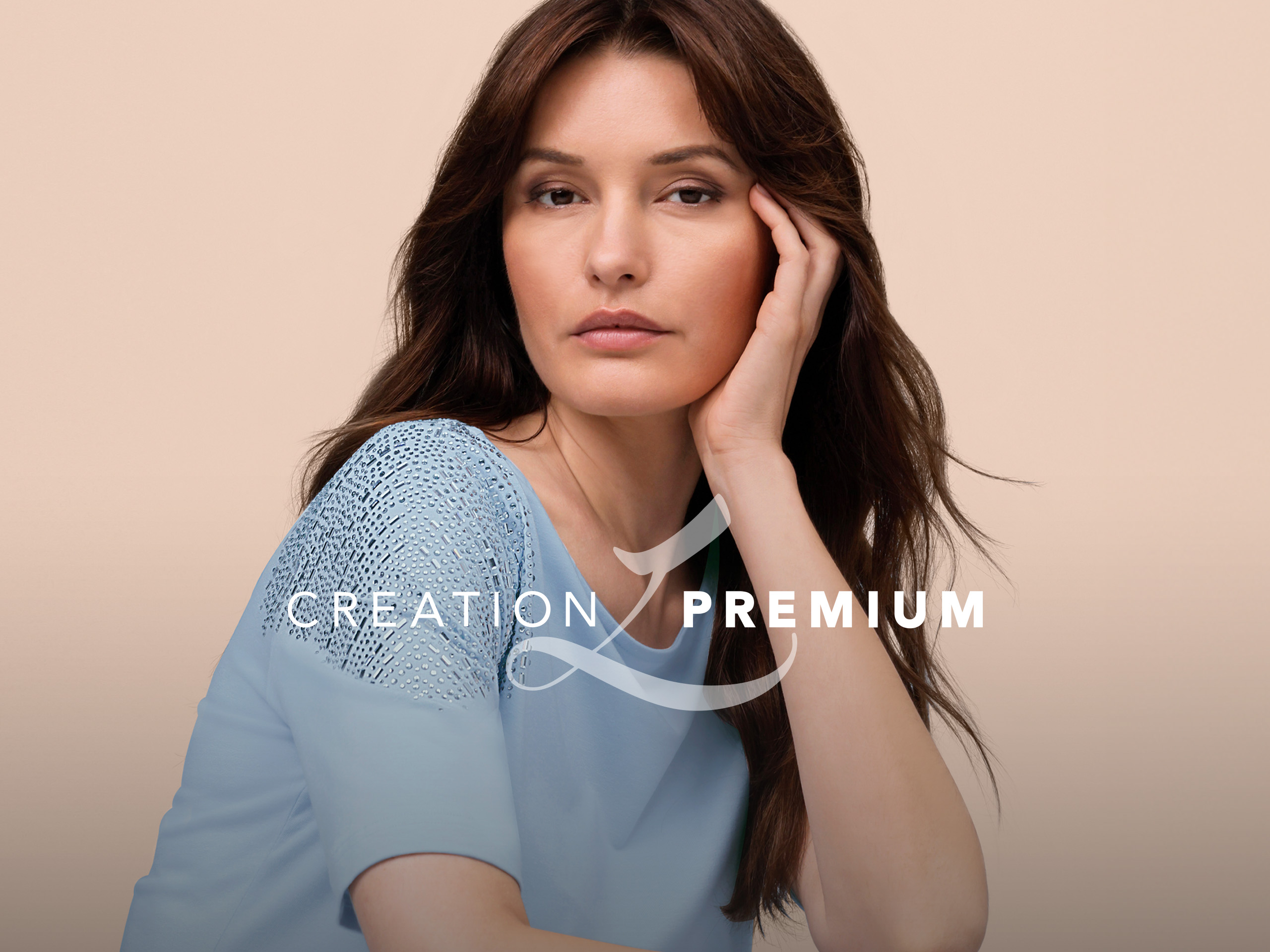 Ein Model trägt ein hellblaues Shirt mit Spitzenbesatz an den Schultern, sitzt vor beigem Hintergrund, rechts CREATION PREMIUM Logo.