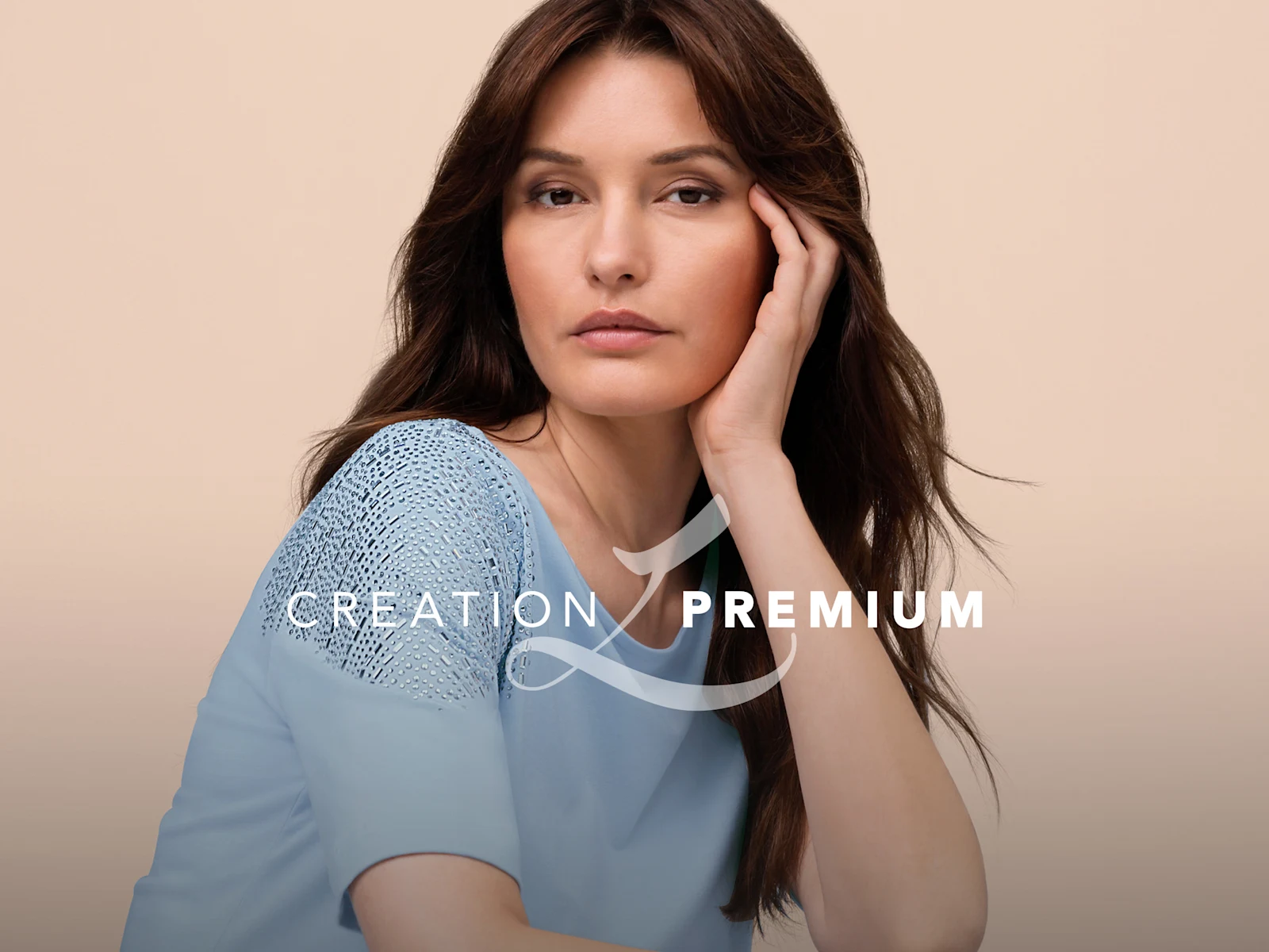 Ein Model trägt ein hellblaues Shirt mit Spitzenbesatz an den Schultern, sitzt vor beigem Hintergrund, rechts CREATION PREMIUM Logo.