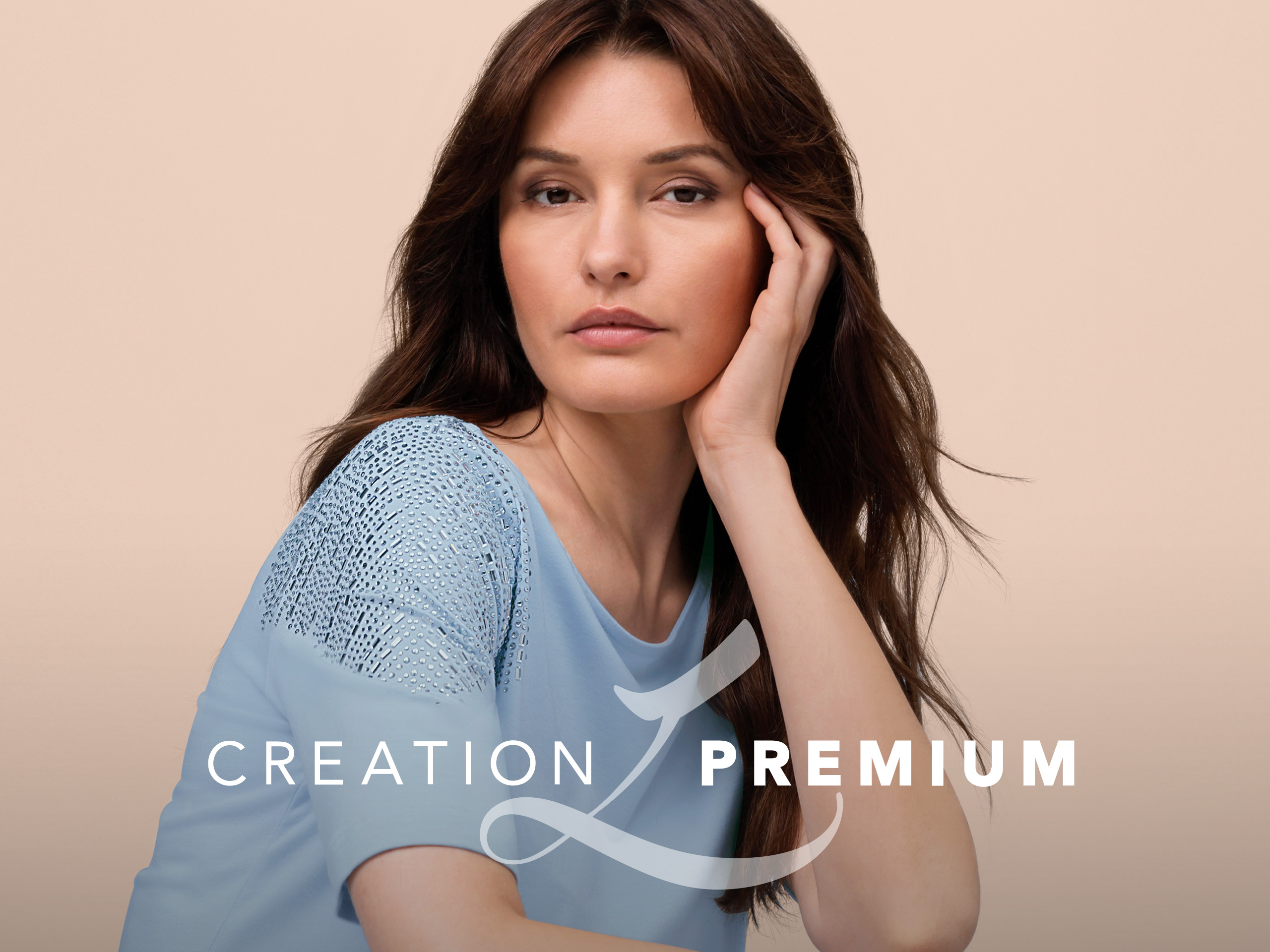 Ein Model trägt ein hellblaues Shirt mit Spitzenbesatz an den Schultern, sitzt vor beigem Hintergrund, rechts CREATION PREMIUM Logo.