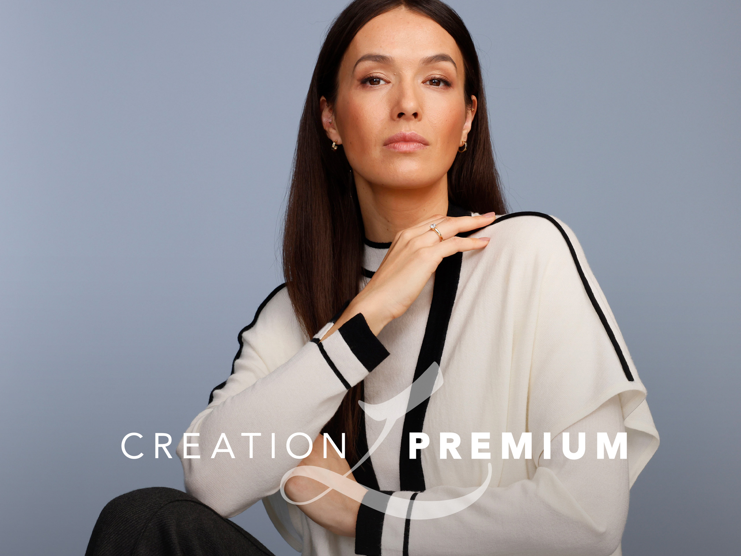 Ein Model trägt einen cremefarbenen Pullover mit schwarzen Streifen, sitzt vor graublauem Hintergrund. Text: 'Creation Premium'.