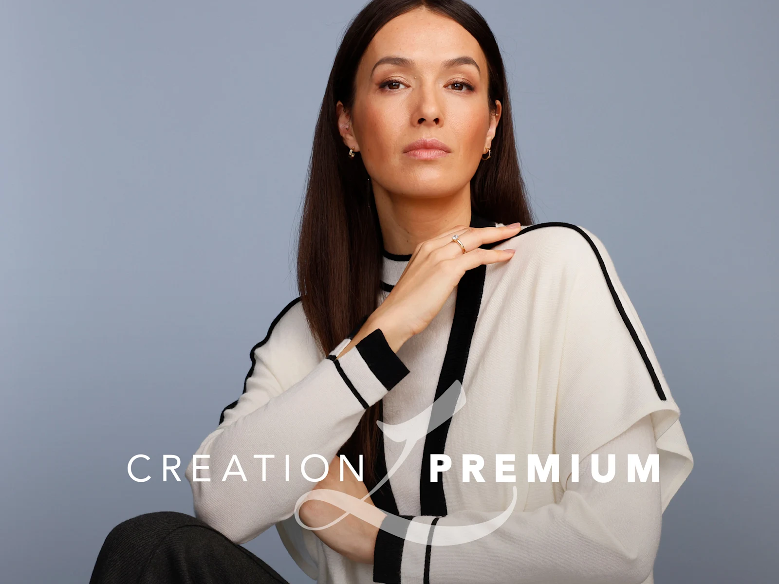 Ein Model trägt einen cremefarbenen Pullover mit schwarzen Streifen, sitzt vor graublauem Hintergrund. Text: 'Creation Premium'.