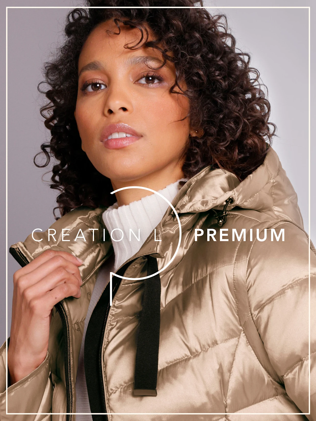 Ein Model mit dunklen Locken trägt eine beige, glänzende Steppjacke mit Kapuze. Das Logo lautet  'CREATION L PREMIUM'. Der Hintergrund ist grau.