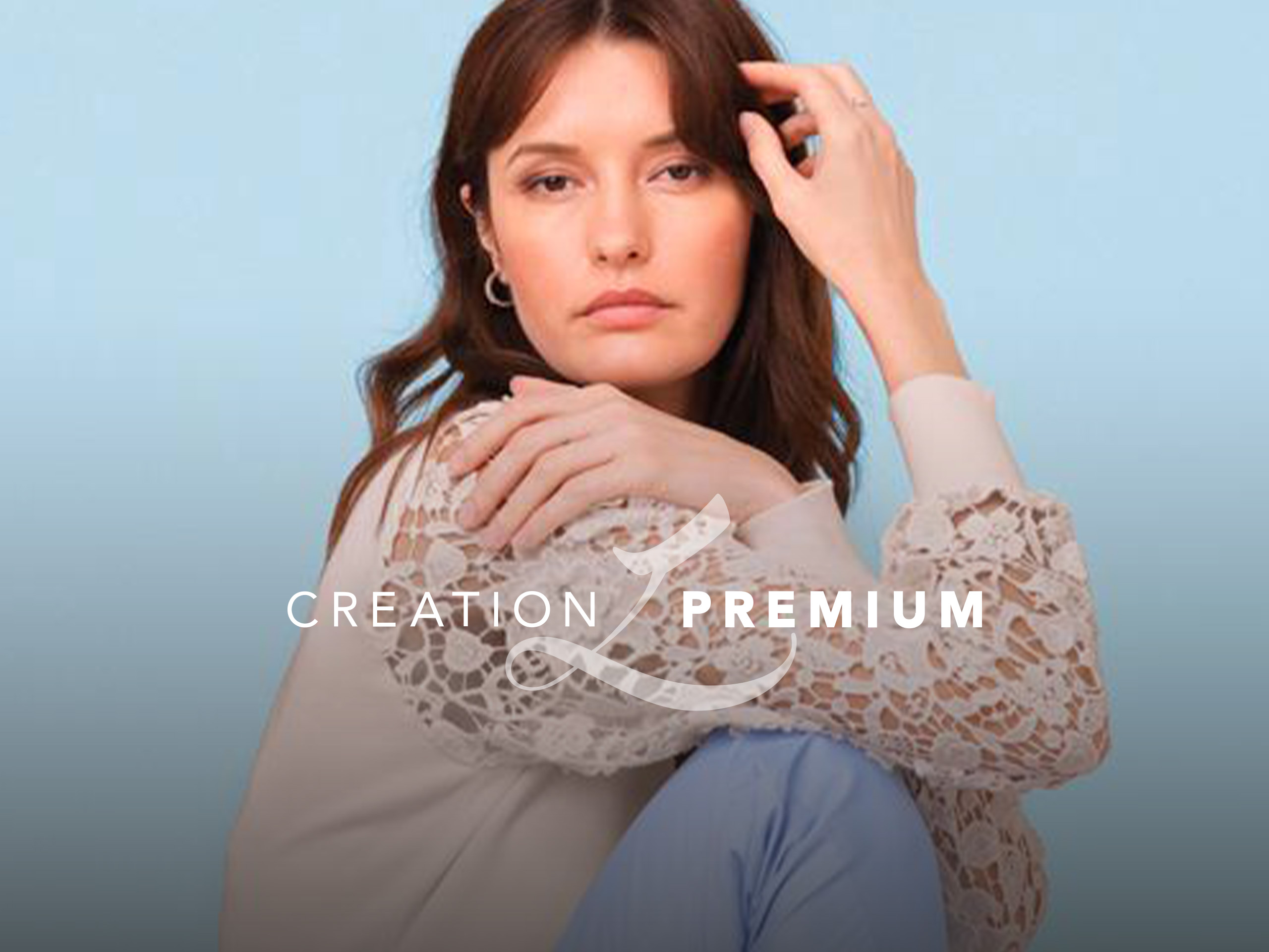 Ein Model sitzt vor hellblauem Hintergrund. Es trägt eine cremefarbene Bluse mit Spitzenärmeln und eine hellblaue Hose. Text: 'CREATION PREMIUM'.